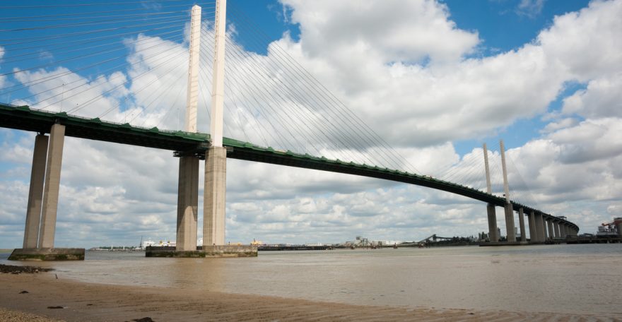 Queen Elisabeth II bridge over river Thames in Dartford with pebbles in the foreground against blue sky with white clouds